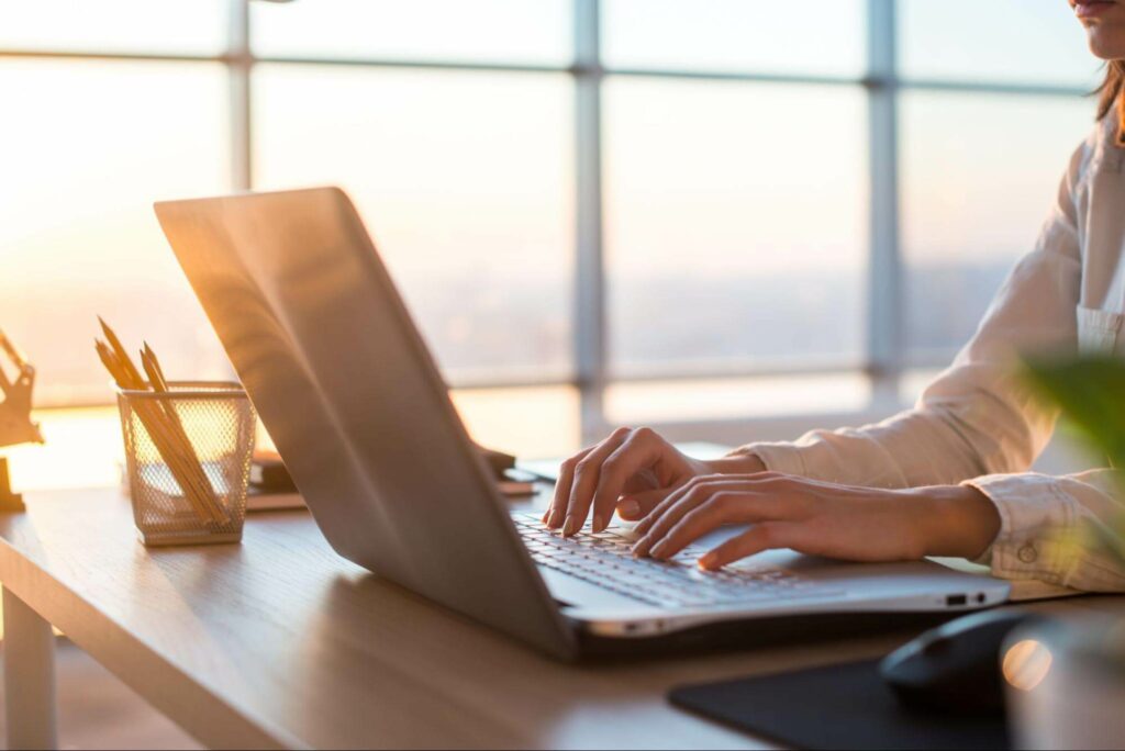 A woman writes a blog on a laptop while sitting in front of a wall of windows at sunset.