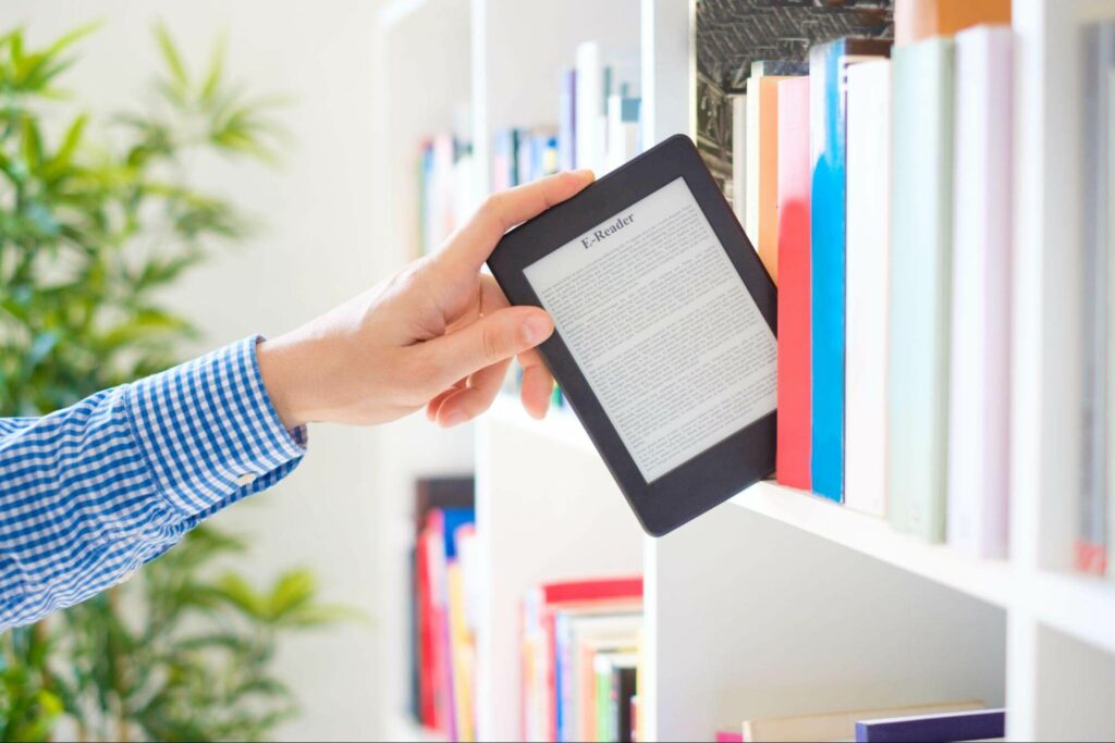 A man in a long-sleeved shirt pulls an ebook reader off a bookshelf.