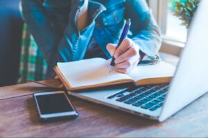 A content marketer rests a hand holding a pen on an open notebook which lies on a laptop keyboard beside a locked smartphone.