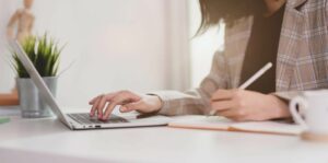 A woman in a brown blazer performs UX copywriting tasks with a notebook and laptop.
